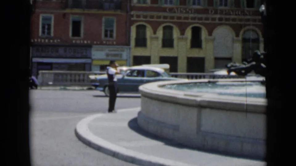 Cannes 1962 street scene with a man near the fountain, classic cars, a two-wheeled vehicle, and CAISSE D'EPARGNE signage in the background.