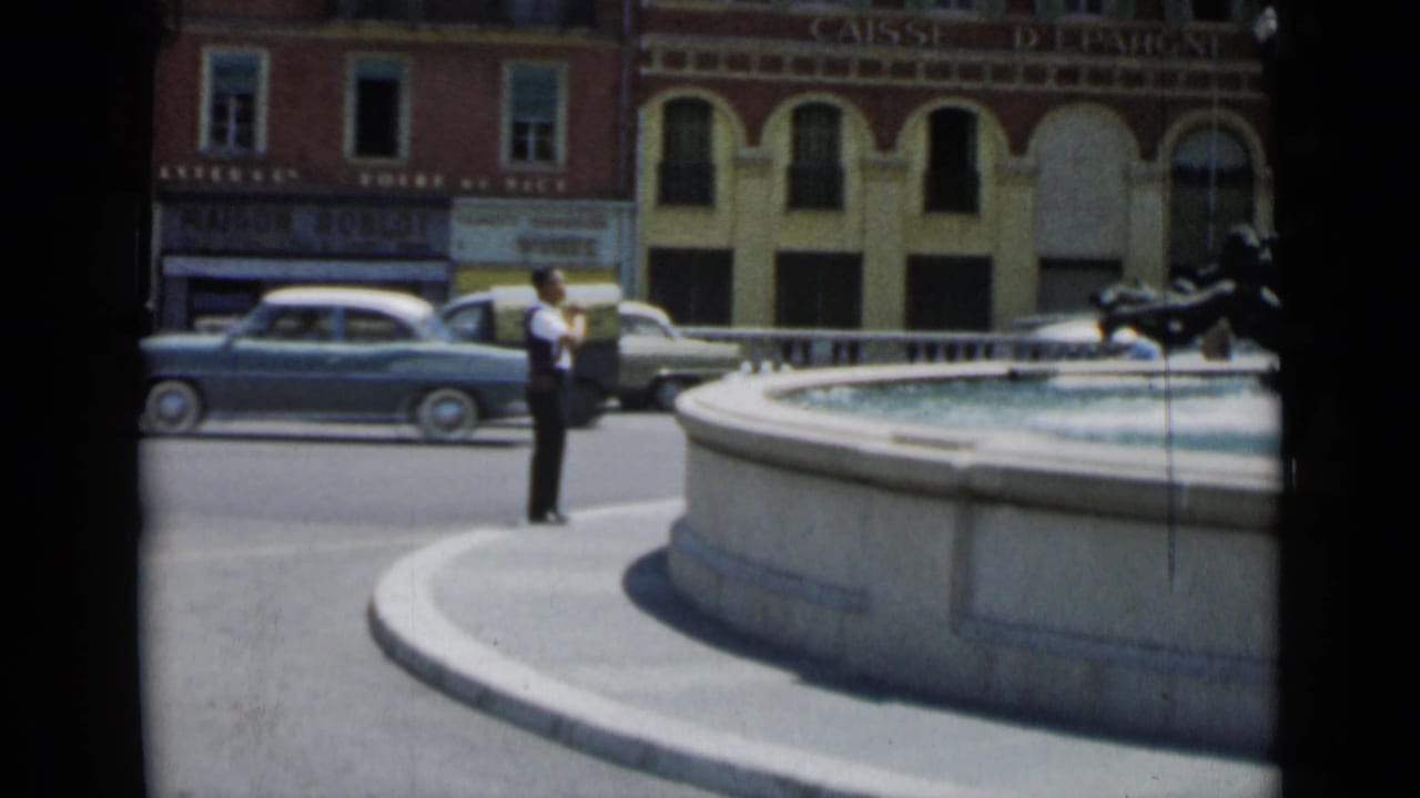 Man in a white shirt stands beside a large fountain as vintage cars pass a building with arched windows.