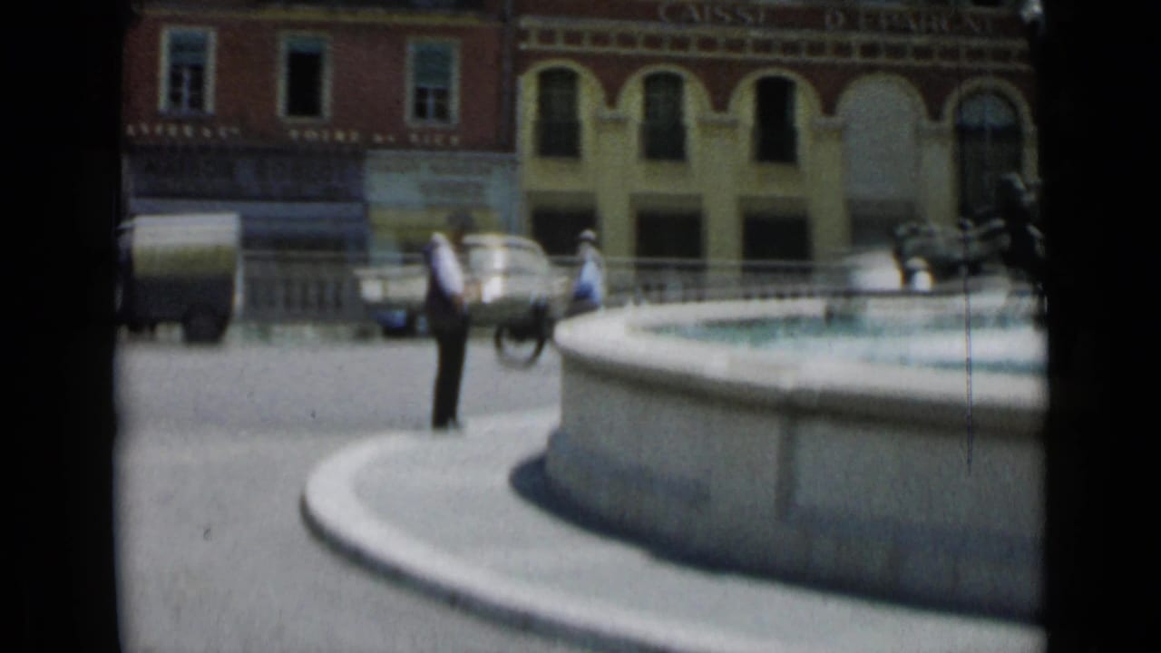 Man remains near the fountain while pedestrians and vehicles move across the square behind him.