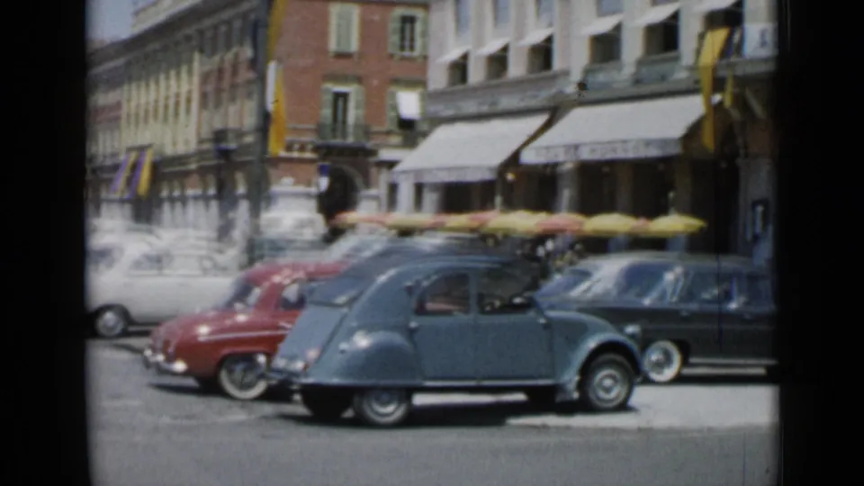 Vintage cars drive through a Cannes commercial street with awnings, pedestrians, and multi-story buildings visible behind them.