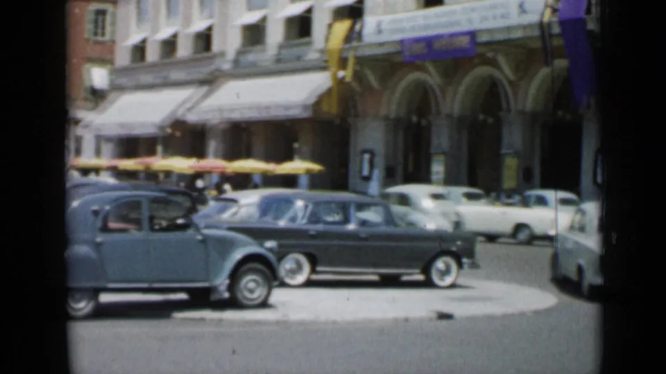 A Cannes street scene shows parked and moving 1960s cars, yellow cafe awnings, arched storefronts, and sunlit building fronts.