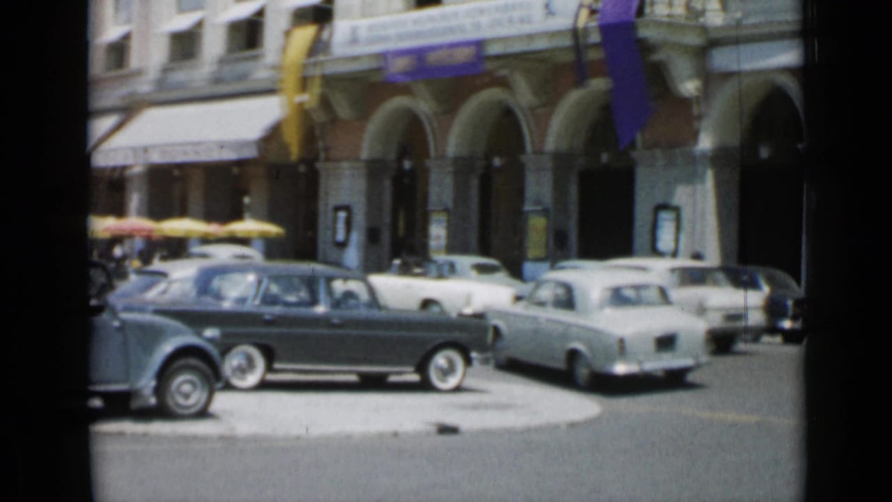 Classic cars on a sunny street in front of an arched building with flags and awnings.