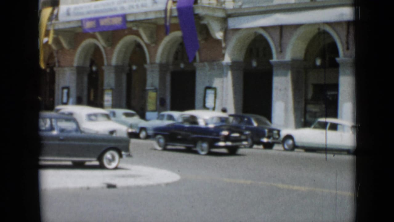 Several classic cars line a street beside an arched building with purple flags.