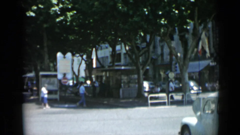 1962 Cannes street view with pedestrians near signs and storefronts as a light-colored car enters the right edge of the frame.