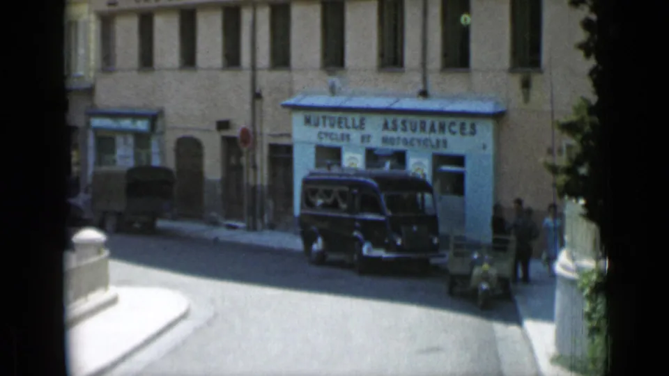 Cannes street corner in 1962 showing a French insurance storefront, parked dark van, pedestrians, and pale sunlit pavement.