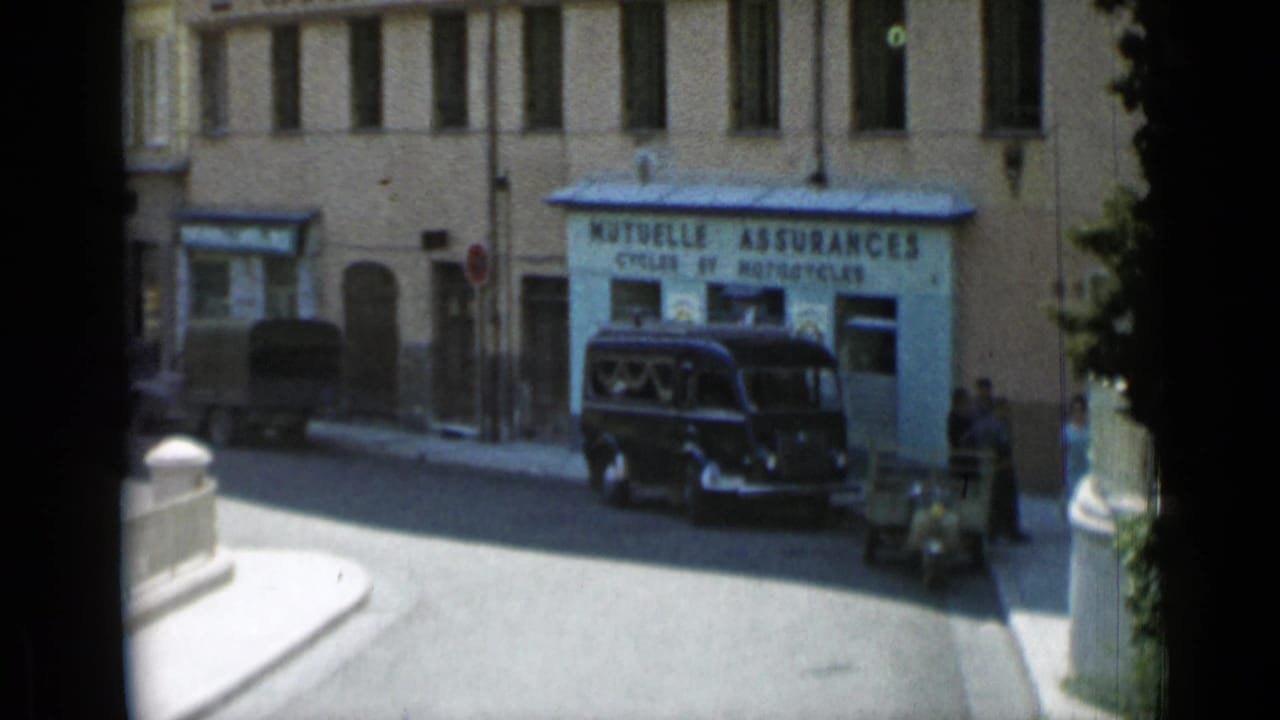 Street corner with storefront signage, a dark vintage car, and pedestrians near tan buildings.