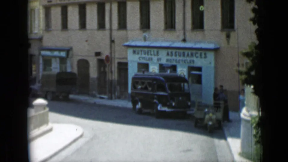 1962 Cannes street view with tan buildings, a storefront sign, a dark vintage car, and pedestrians in bright daylight.