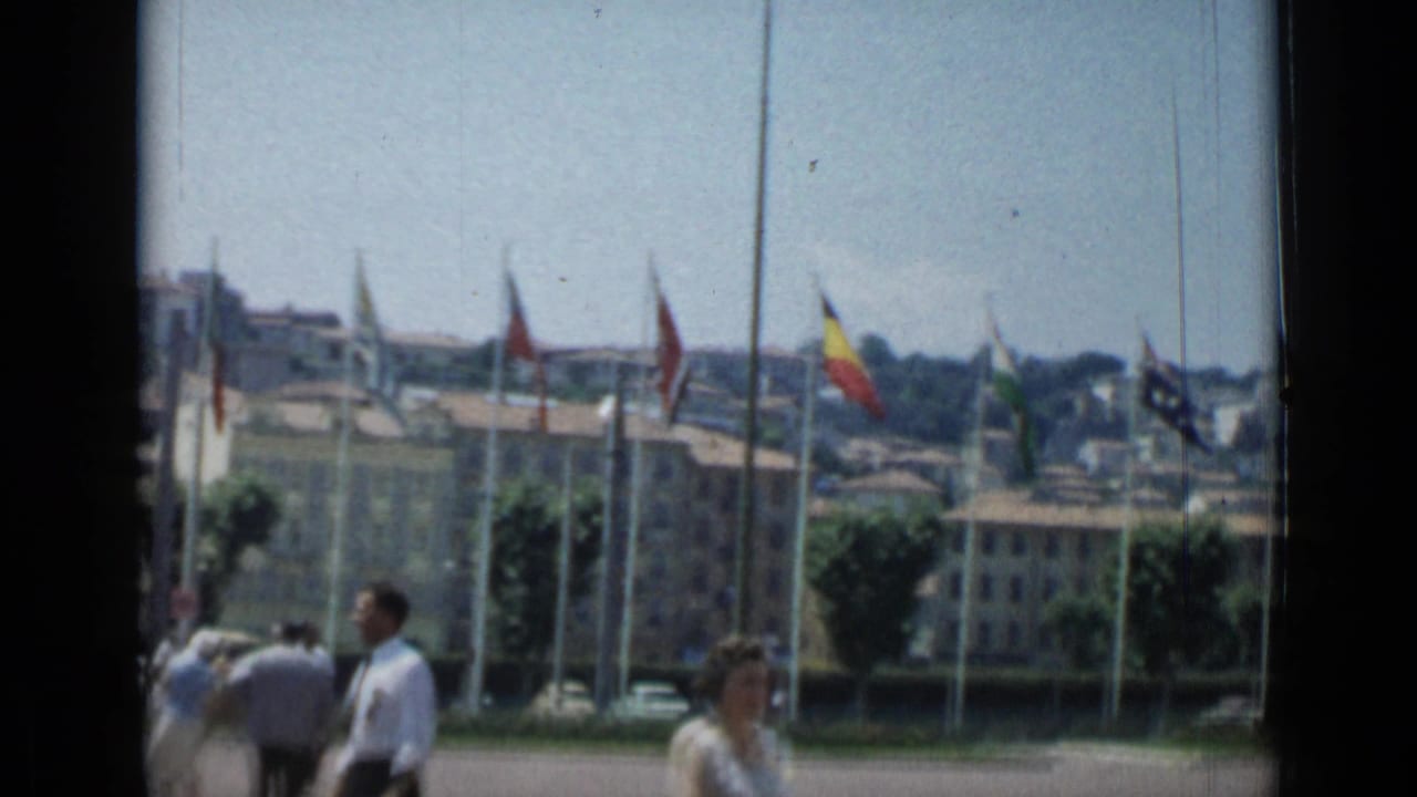 Cannes public area with international flags flying before low buildings and distant hillside structures in 1962 home-movie footage.