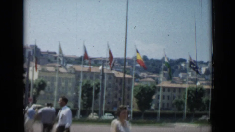 Cannes public area with international flags flying before low buildings and distant hillside structures in 1962 home-movie footage.