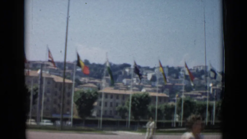 1962 Cannes view of flags on tall poles, tan buildings, pedestrians, trees, and distant hillside structures under bright sky.