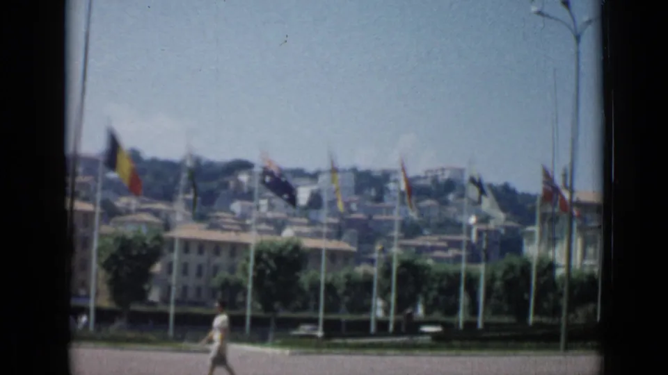 Wide 1962 Cannes home-movie frame showing international flags, tan buildings, walkers, trees, and hillside structures in sunlight.