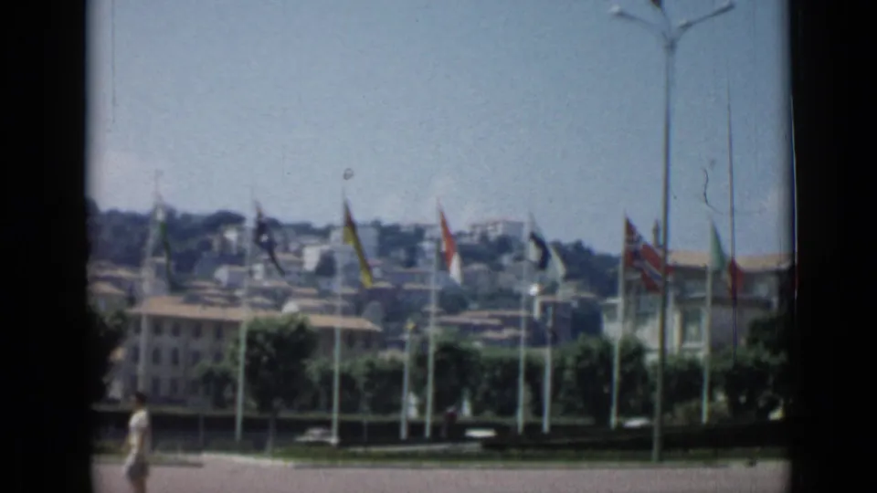 Cannes, France, 1962 home-movie frame showing rows of flags, hillside buildings, trees, and a pedestrian crossing the foreground.