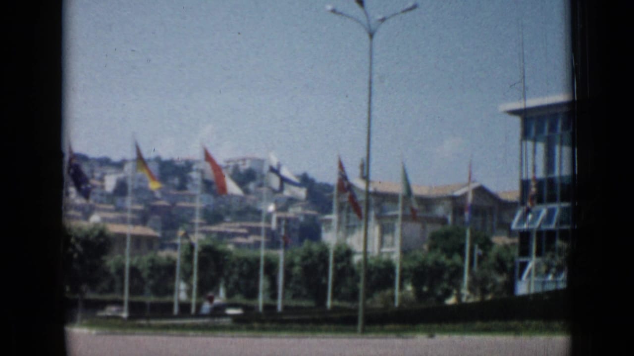 Flags and a modern glass building with hillside structures in the background.