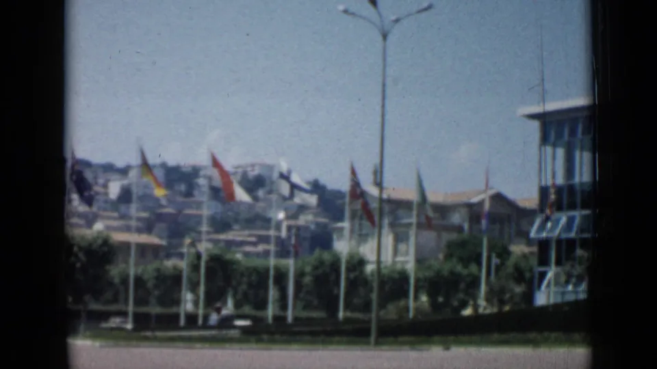 Cannes, France, 1962 frame with national flags, a modern building edge, hillside rooftops, trees, and a car near the road.