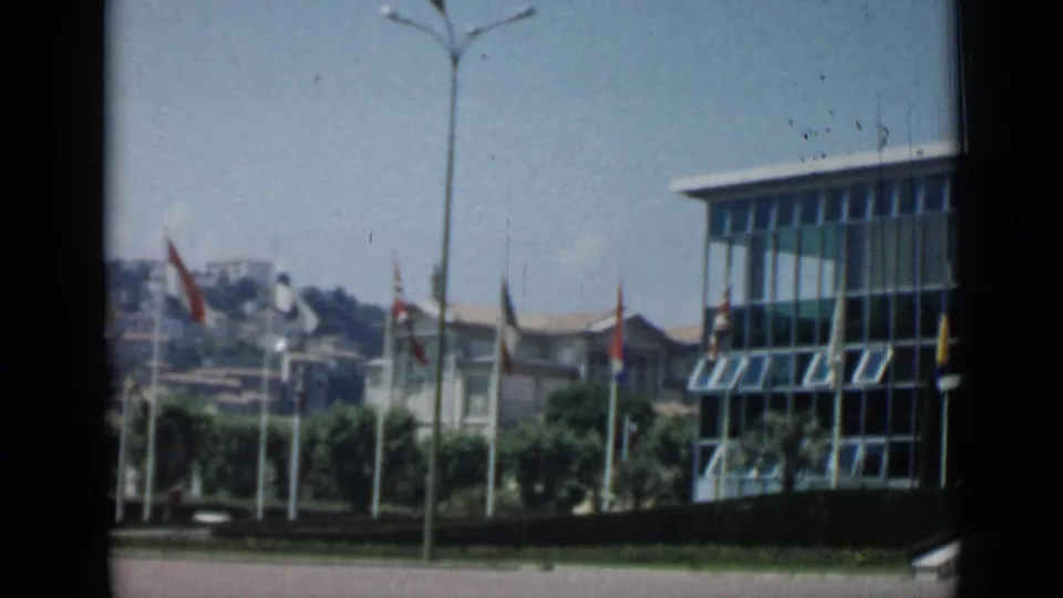 Cannes, France, 1962 home-movie frame showing a glass-fronted building, flags, trees, streetlight, and hillside buildings.