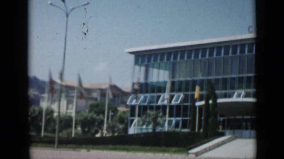 Cannes, France, 1962 frame focused on a modern glass building with open windows, nearby flags, trees, and a paved approach.