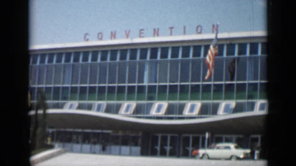 Closer 1962 Cannes view of the convention facade, flags, open windows, entrance area, and a light-colored car near the curb.