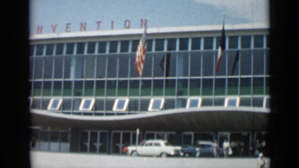 1962 Cannes convention building with flags, classic cars, entrance doors, and small pedestrians visible near the curb.