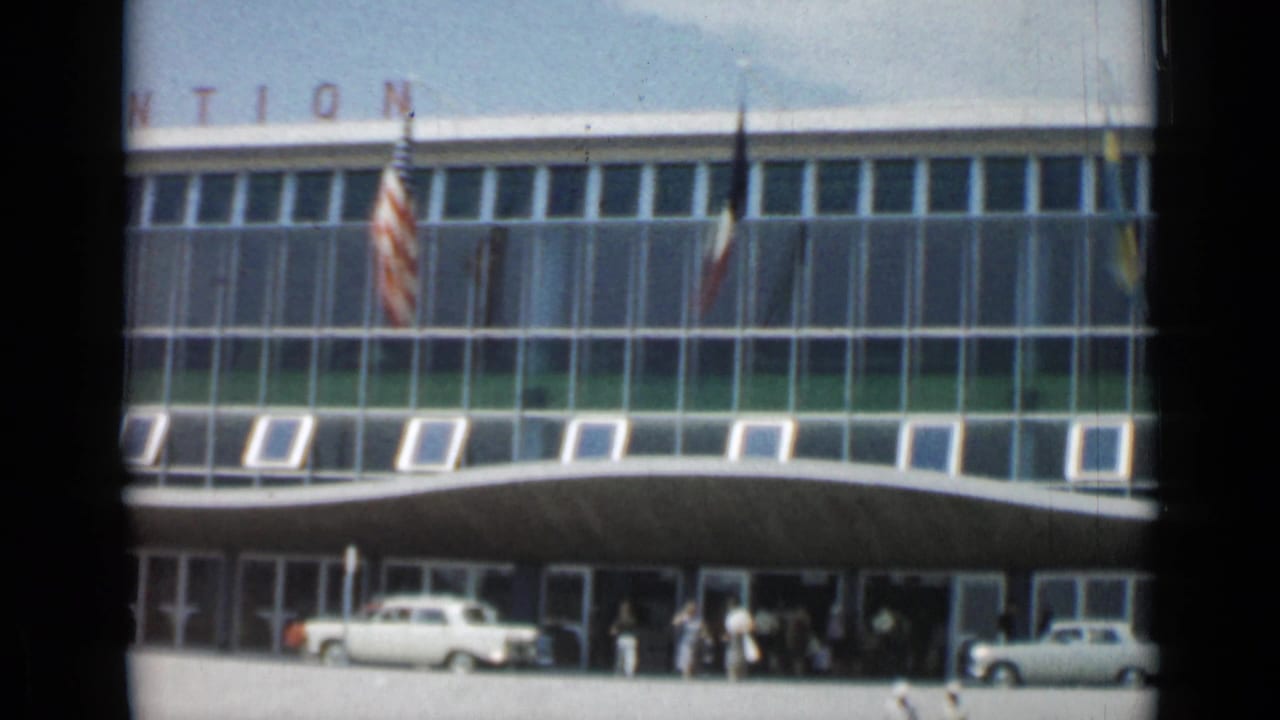 Glass-fronted building entrance with flags, cars and people near the doors.