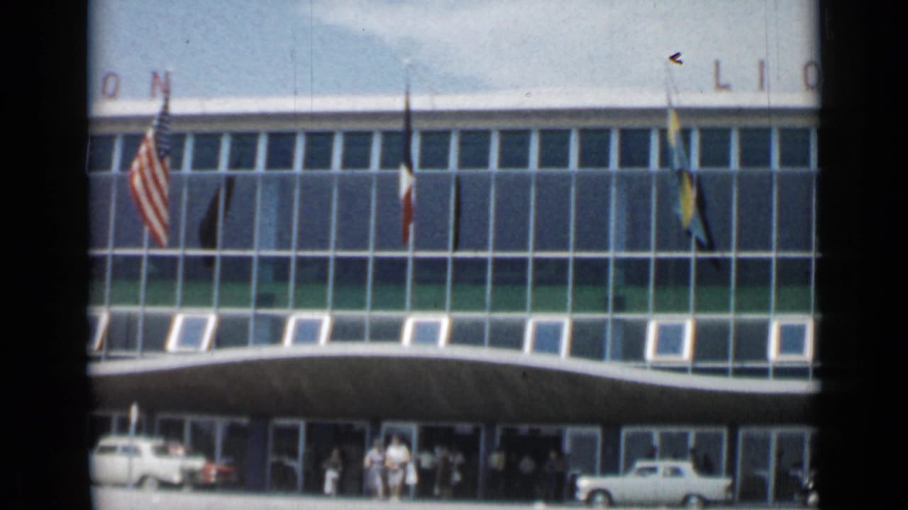 Building facade with flags, cars and people entering near the doors.