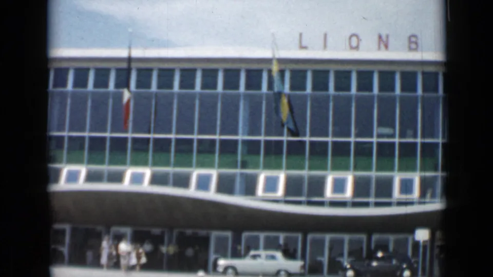 1962 Cannes frame showing the building's glass grid, flags, curved canopy and visitors near a white car.