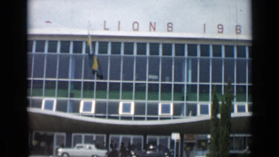 1962 Cannes home-movie frame with Lions roofline lettering, flags, cars and pedestrians at the glass-fronted entrance.