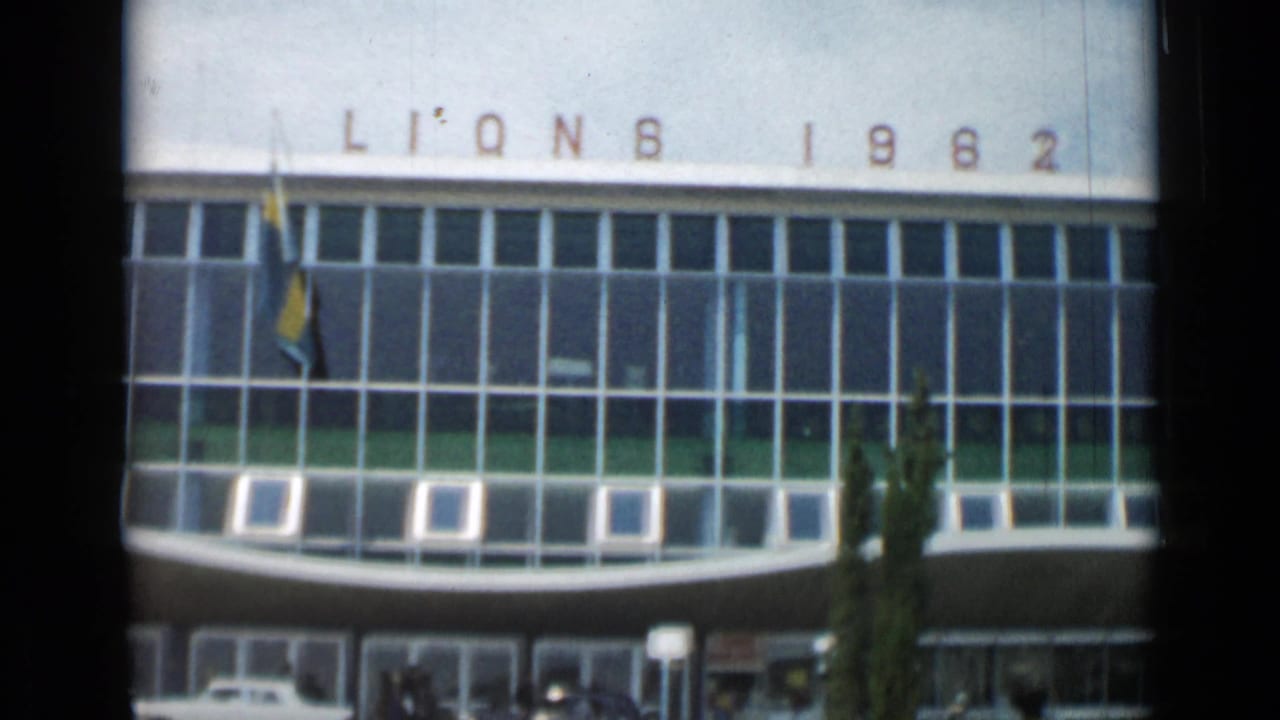 1962 Cannes 8mm frame showing a modern glass-fronted building with partial “LIONS 1962” facade lettering, a flagpole, cypress tree, entrance canopy, and parked cars near the doorway.