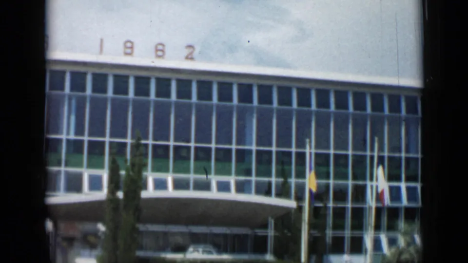 1962 Cannes 8mm frame showing the camera pan across the building facade, with visible “1962” numerals, a cypress tree, multiple flags, the curved canopy, and cars near the entrance.