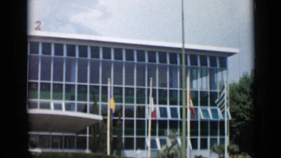 Final 1962 Cannes frame from the pan, showing the right side of the modern glass facade with flagpoles, several flags, open awning windows, trees, and a partly cloudy sky.
