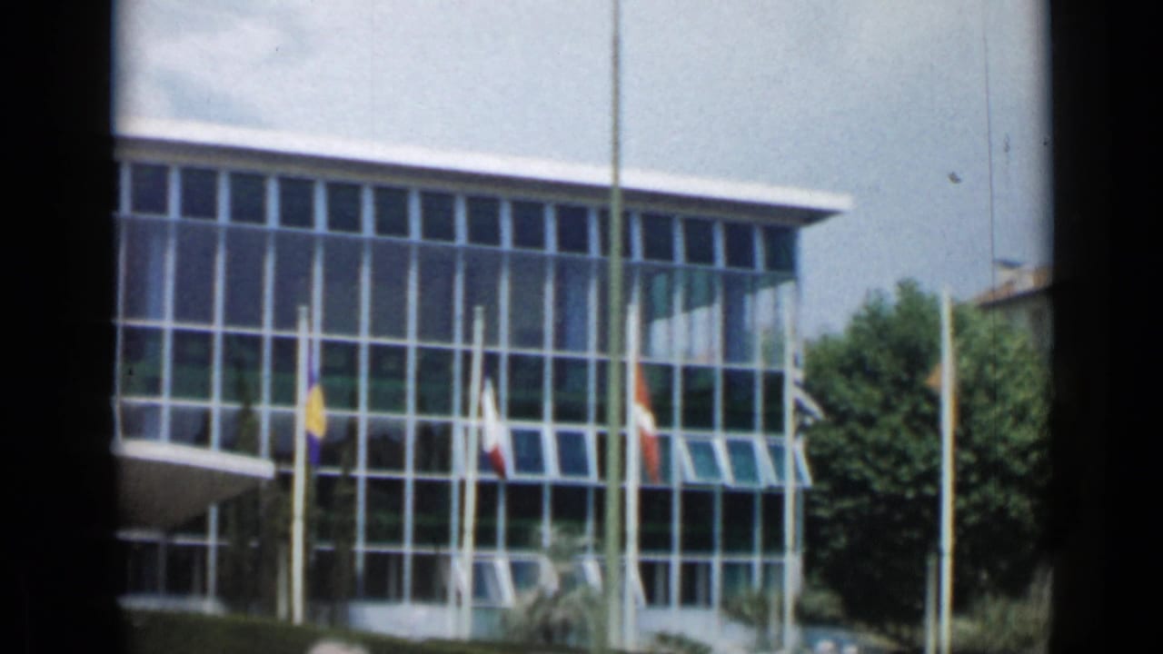 White modern building with large glass windows and several flagpoles in front.