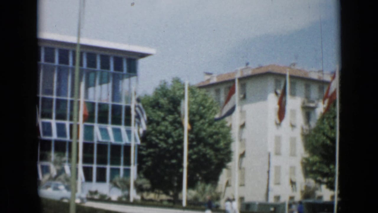 Cannes, 1962 view of a glass-fronted white building beside trees, flagpoles, and a pale multi-story building.