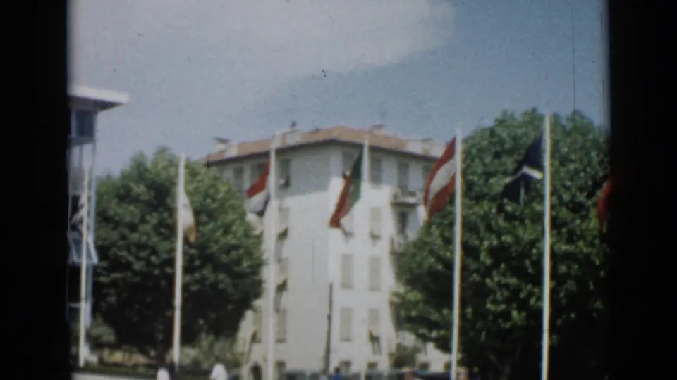 1962 Cannes home-movie frame with a pale multi-story building, leafy trees, and flags waving on tall poles.