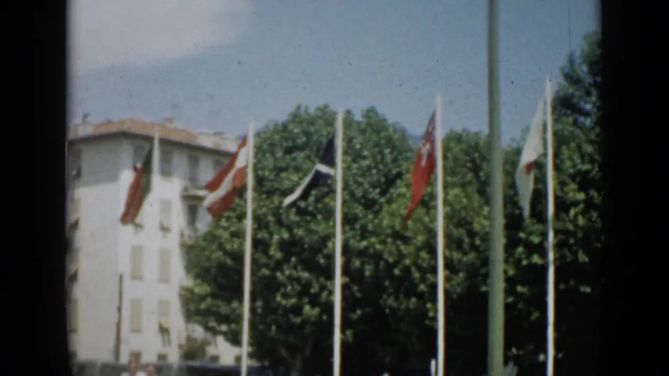 Cannes, 1962 frame showing multiple flags on tall poles, leafy trees, and a pale building in bright daylight.