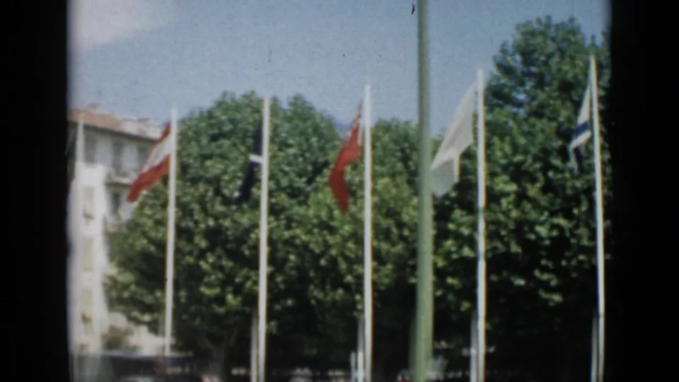 1962 Cannes 8mm frame showing national flags on poles, leafy trees, blue sky, and a pale building at the left edge.