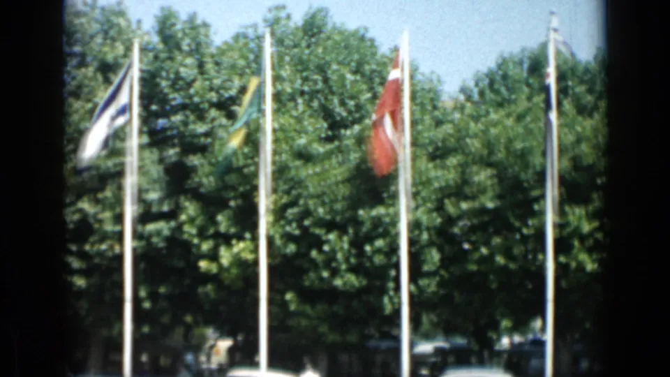 1962 Cannes 8mm frame showing national flags before leafy trees, with partial parked cars and deep frame borders.