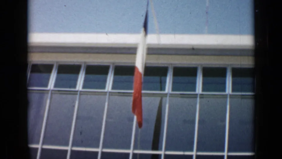 French flag hanging before a modern windowed building in Cannes, France, from 1962 8mm footage.