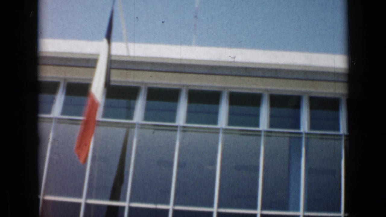 French tricolor seen against a glass-fronted building facade in Cannes, France, in early-1960s home-movie footage.