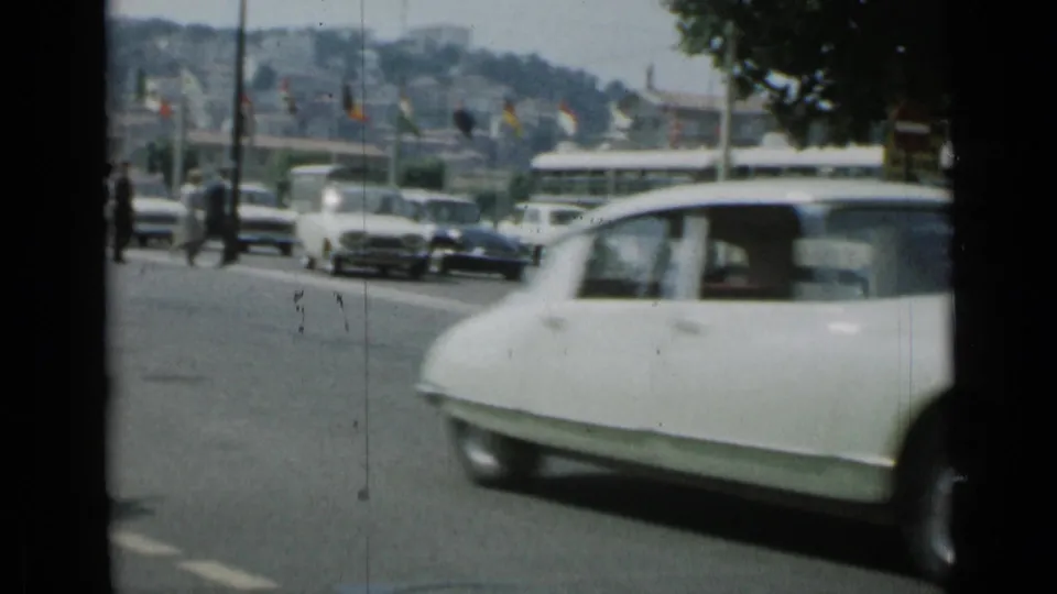 A white Citroen DS-style car passes through a Cannes roadway with buses, cars, and flags in the background.