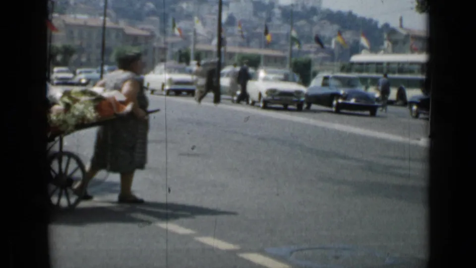A woman with a loaded cart waits beside a Cannes street while cars and pedestrians move through the background.