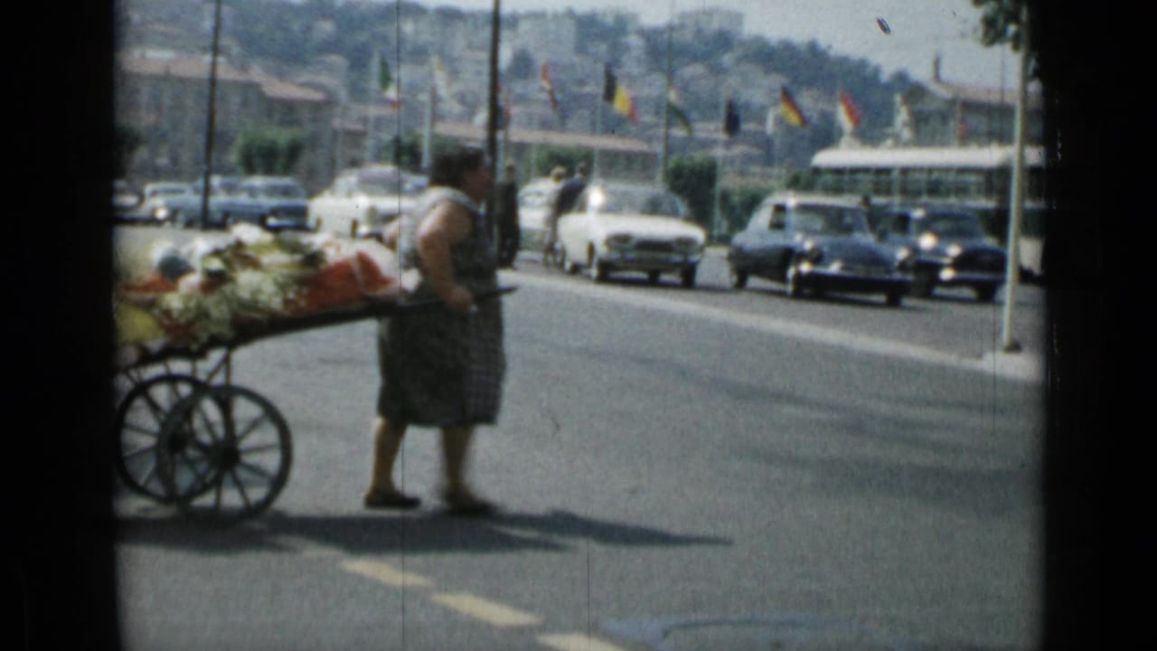 Woman pulling a loaded produce cart across a busy street with cars and flags behind her.