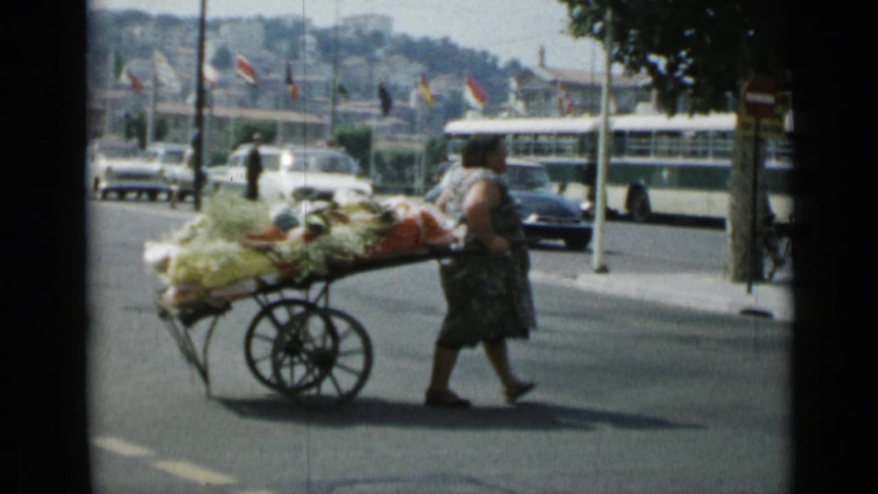 The woman continues pulling the loaded produce cart through Cannes traffic, with vintage cars, street flags, and a green-and-white bus behind her.