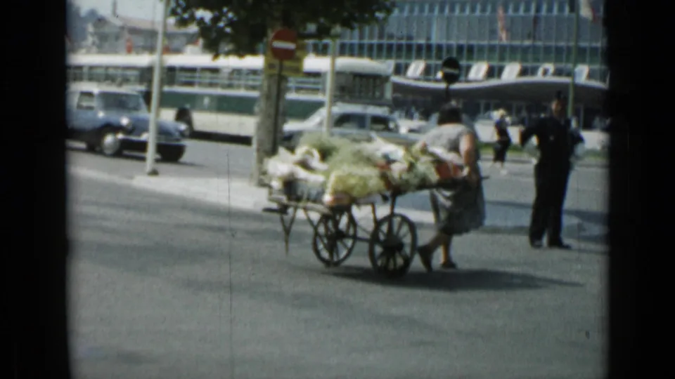 The woman pauses or turns the produce cart near the curb in Cannes, with pedestrians, a bus, cars, and street signs visible nearby.