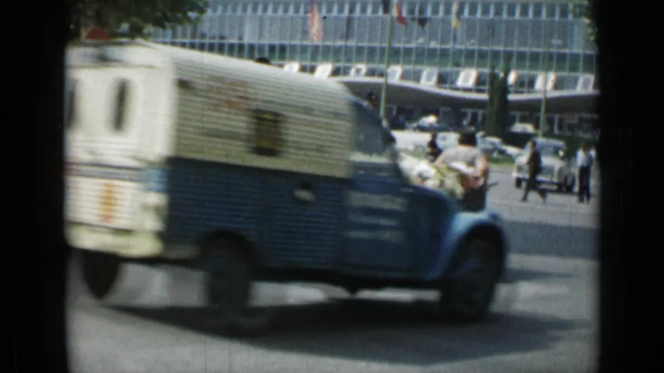 A blue-and-white van moves through a Cannes street in 1962, with pedestrians and cars near a modern facade.