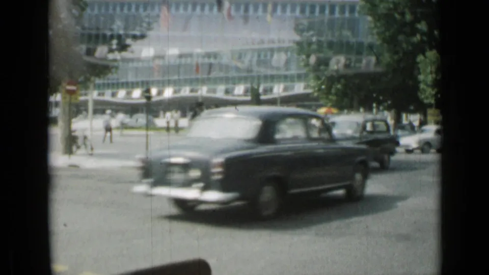 A dark classic car moves across a 1962 Cannes street as pedestrians cross near a modern building and road signs.