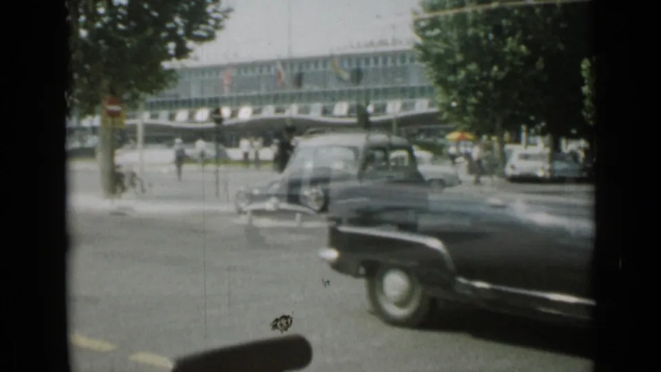 A woman pushes a loaded two-wheeled cart as a classic car passes on a Cannes street in 1962.