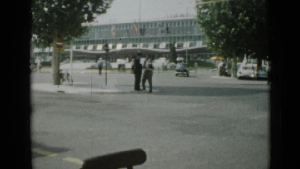 1962 Cannes street view from a vehicle, showing pedestrians near an intersection, classic cars, trees, and a modern glass-fronted building.