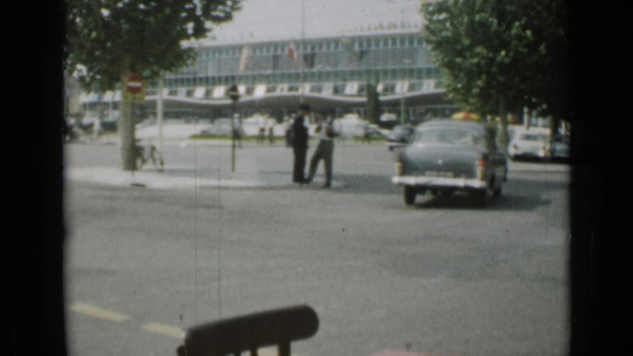 A dark classic car moves through the 1962 Cannes intersection while pedestrians wait near trees and a large glass-fronted building.