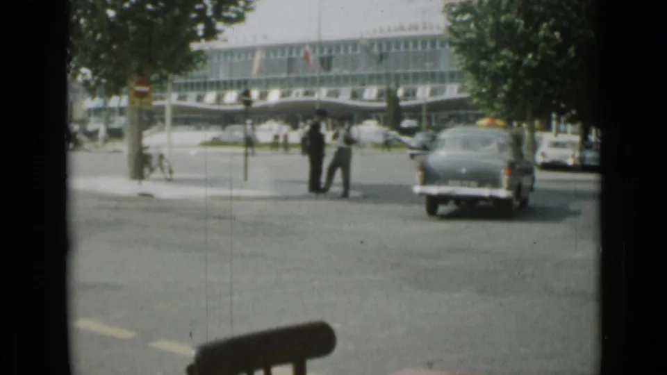 A dark classic car moves through the 1962 Cannes intersection while pedestrians wait near trees and a large glass-fronted building.