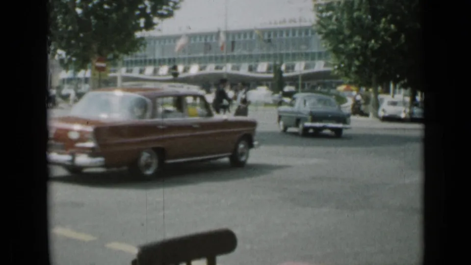 A red sedan crosses the foreground in 1962 Cannes, with another car, pedestrians, street trees, and a modern building beyond.