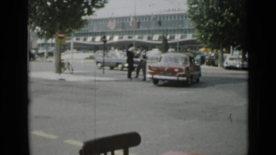 The 1962 Cannes intersection remains busy as a red car passes pedestrians, trees, parked vehicles, and a glass-fronted building.